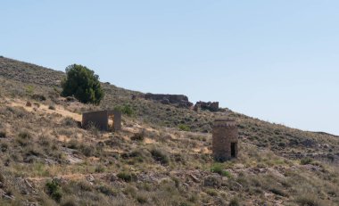 Eski ve terk edilmiş bir maden kompleksinde binalar tuğlalarla örülmüş, taş bir duvar var, dağlık bir alan ve gökyüzü açık.