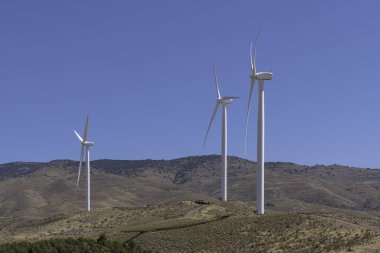 Güney İspanya 'daki dağlarda rüzgar türbinleri, ağaçlar ve kuru otlar var, bir elektrik hattı çalışıyor ve gökyüzü temiz.