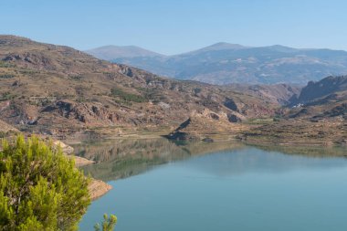 Güney İspanya 'da Beninar Barajı, suda yansımalar var, çam ağaçları ve çalılar var, gökyüzü açık.