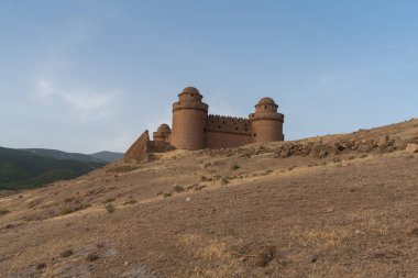 Güney İspanya 'daki bir dağın tepesindeki La Calahorra şatosu, kale taştan yapılmış, 3 kulesi var, çimenler kuru, gökyüzü bulutlu.