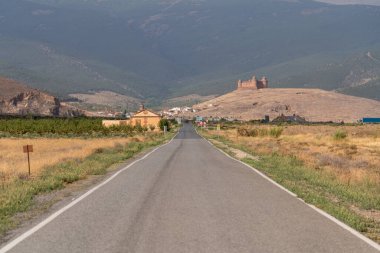 La Calahorra kalesine giden yol, bir ev ve bir kasaba, dağlar, ağaçlar ve çimenler var.