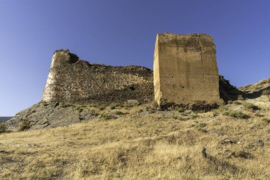 Eski bir kalenin eski duvarı, duvar taş ve çamurdan yapılmıştır, kayalık bir alandır ve çimenler vardır, gökyüzü açıktır.