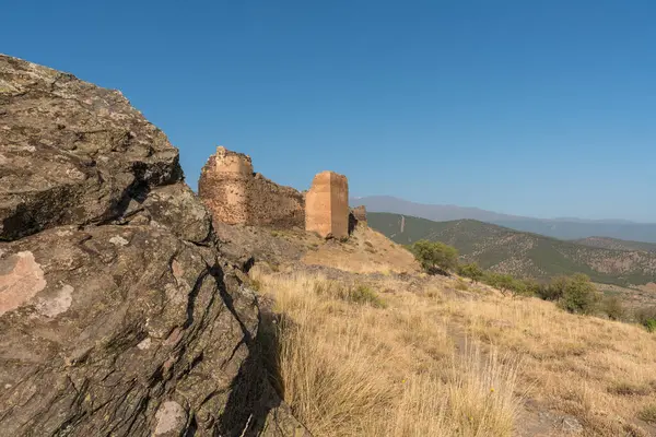 Eski bir kalenin eski duvarı, duvar taş ve çamurdan yapılmıştır, kayalık bir alandır ve çimenler vardır, gökyüzü açıktır.