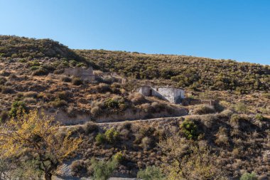 Güney İspanya 'da La Alpujarra' nın dağlık arazisi, ağaçlar ve çalılar var, harabeler içinde bir çiftlik evi var, gökyüzü açık.