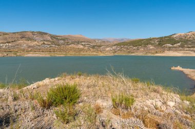 Güney İspanya 'daki dağların arasında Beninar rezervuarı var. Çalıların ve ağaçların bitkileri var. Gökyüzü açık.