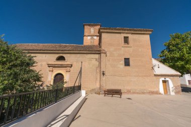 Güney İspanya 'da küçük bir kasabanın kilisesi, kilisenin bir kulesi var ve tuğladan yapılmış, birden fazla kapı ve pencere var, 2 ahşap bank var, ağaçlar var.