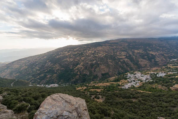 Güney İspanya 'da Sierra Nevada' da dağlık bir arazi, dağın eteklerinde bir köy var, çam ormanları ve çalılar var, gökyüzünde bulutlar var.