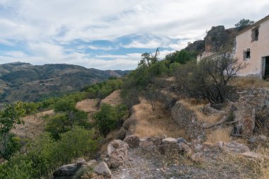 Güney İspanya 'nın dağlık arazisi, birkaç taş duvar, harabe bir çiftlik evi, ağaçlar, çalılar ve kuru otlar var, gökyüzünde bulutlar var.