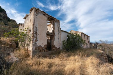 Güney İspanya 'daki harabe çiftlik evi, taştan yapılmış, birkaç pencere ve bir kapı, dağlar, ağaçlar, çalılar ve kuru çimenler var, gökyüzünde bulutlar var.