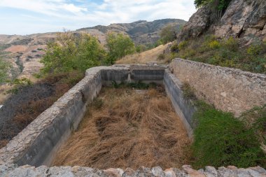 Dağlarda eski bir havuz, taş ve betondan yapılmış bir havuz, ağaçlar ve bol bitki örtüsü, gökyüzünde bulutlar var.