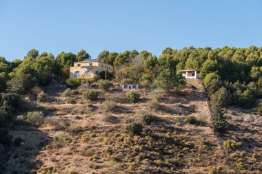 Dağda birkaç çiftlik evi, ağaçlar, çalılar ve çimenler var, gökyüzü açık.