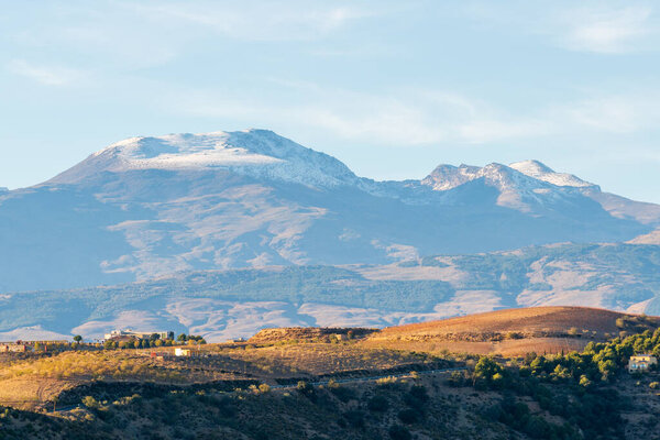 mountainous landscape in southern Spain, there are trees on the mountain, there is a farmhouse, on the top of the mountain there is snow, the sky has clouds