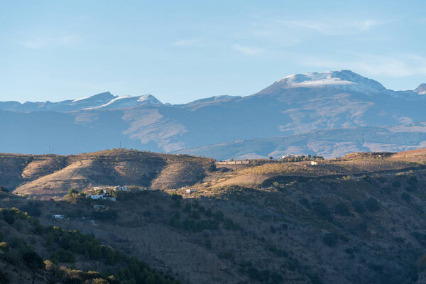 mountainous landscape in southern Spain, there are trees on the mountain, there is a farmhouse, on the top of the mountain there is snow, the sky has clouds