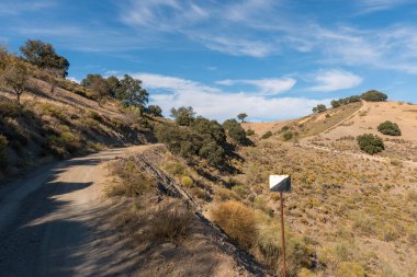 Güney İspanya 'nın dağlık arazisi, eski bir metal işareti var, dağda ağaçlar ve çalılar var, orman yolu var, gökyüzünde bulutlar var.