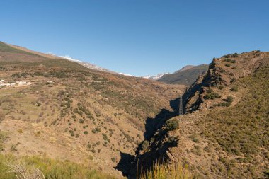 Sierra Nevada 'nın dağlık arazisinde ağaçlar, çalılar ve çimenler var. Dağ yamacında bir kasaba var. Dağların arasından bir nehir geçiyor.