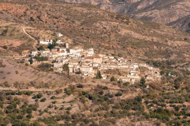 Small town on the side of a mountain in southern Spain, its houses have white painted facades, there is a church, there are trees and bushes