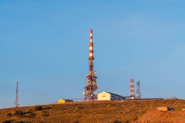 Telekomünikasyon antenleri dağda, çeşitli binalar, bitki örtüsü ve taşlar var, gökyüzü açık.