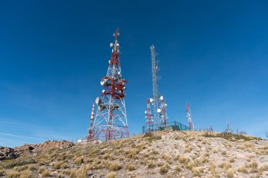 Telekomünikasyon antenleri dağda, çeşitli binalar, bitki örtüsü ve taşlar var, gökyüzü açık.