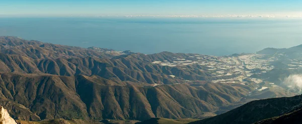 mountainous area in the south of Spain, there are greenhouses in the valley, there are clouds, in the background is the Mediterranean Sea