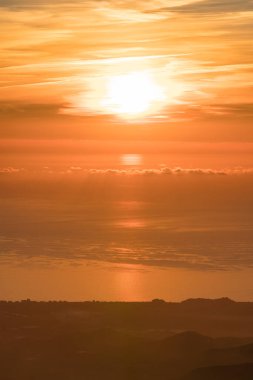 Akdeniz kıyısında gün batımı, dağda bitki örtüsü, sahilde evler, gökyüzünde bulutlar var.