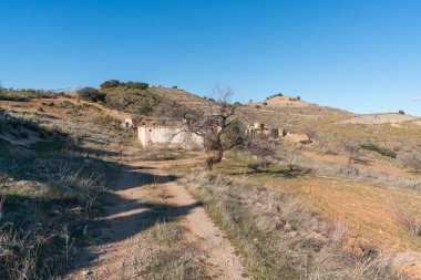 Dağdaki toprak yolda, harabe bir çiftlik evi var, ağaçların ve çalıların bitkileri var, taşlar var, gökyüzü açık.