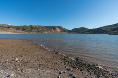 Güney İspanya 'da Beninar rezervuarı, bitki örtüsü olan dağlar, kıyıda çamur ve taşlar var, gökyüzü açık.