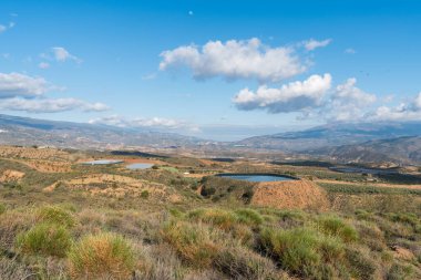 Güney İspanya 'nın dağlık arazisi, bir su deposu var, seralar var, ağaçlar ve çalılar var, gökyüzü bulutlu.