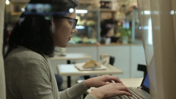 Ela trabalha com o computador, vista através da janela de um café. Trabalho de escritório à noite — Vídeo de Stock