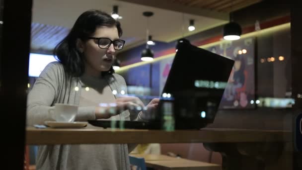 彼女は素敵なカフェでコンピューターで動作、彼女はコーヒーの窓からすのテーブルに座っています。カフェで美しいランプ. — ストック動画