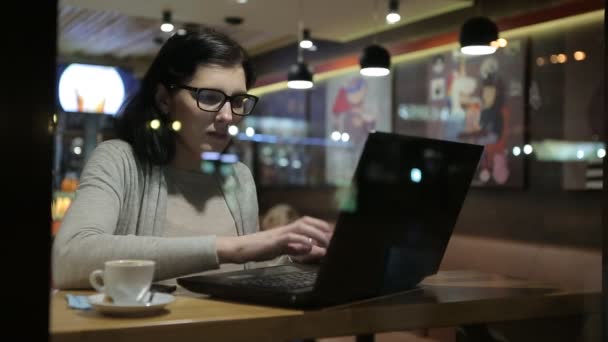 Ela trabalha com computadores em um bom café, ela se senta em uma mesa pelos vidros da janela. Lâmpadas bonitas no café — Vídeo de Stock