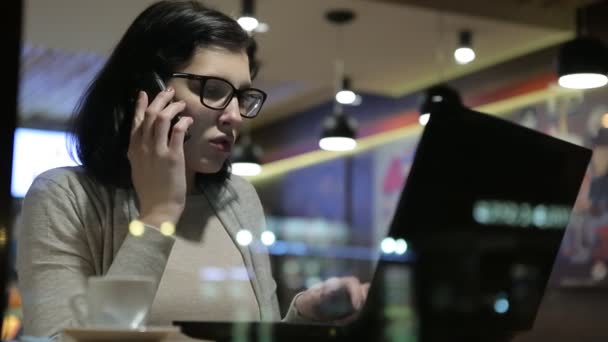 Ela trabalha com computadores em um bom café, ela se senta em uma mesa pelos vidros da janela e fala por telefone . — Vídeo de Stock