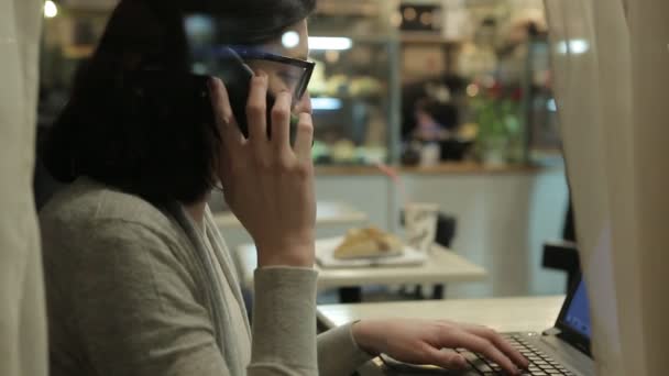 Elle travaille avec l'ordinateur, vue à travers la fenêtre d'un café. Travail de bureau de nuit, elle a dit par téléphone . — Video