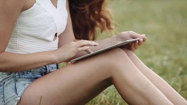 Милая девушка с длинными и красивыми ногами сидя на траве и играя с ноутбуком в руках в медленном движении — стоковое видео