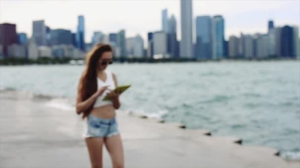 Vacker flicka med långt hår i vit skjorta och jeansshorts är spelar med hennes enhet framför Downtown — Stockvideo