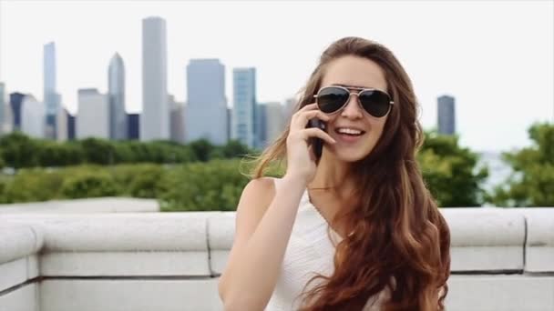 Vackra kaukasiska tjej med långt brunt hår i vit skjorta och svarta solglasögon är talande genom mobiltelefon framför Downtown i Slow Motion — Stockvideo