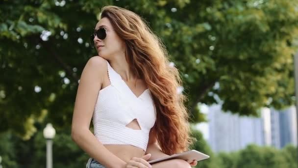 Una hermosa chica sentada en un banco con la tableta en su regazo mirando a su alrededor felizmente con un árbol en el fondo — Vídeos de Stock