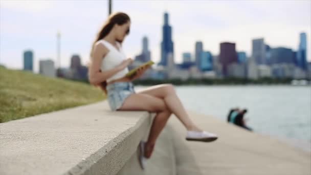 Menina Petite impressionante com cabelos longos está sentado no concreto com seu dispositivo com edifícios altos e lago no fundo em câmera lenta — Vídeo de Stock