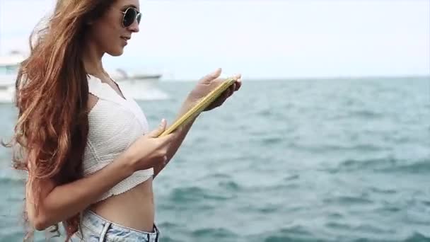Menina bonita com longos cabelos castanhos está andando com seu dispositivo junto ao lago — Vídeo de Stock