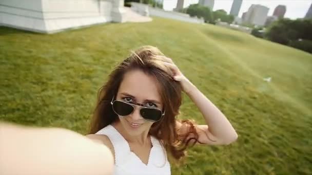 Una chica con el pelo largo marrón y gafas de sol está tomando un video de sí misma en un día soleado brillante en el parque . — Vídeo de stock