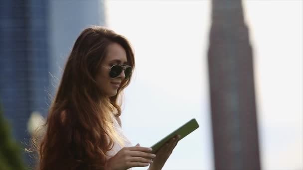 Una chica con el pelo largo y castaño vistiendo una camisa blanca y gafas de sol negras está usando su tableta en la ciudad en un día soleado — Vídeo de stock