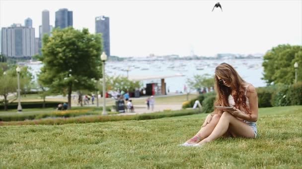 Ragazza carina in camicia bianca e pantaloncini Jean in seduta sull'erba con il suo dispositivo e la gente, Lago e alti edifici sullo sfondo — Video Stock