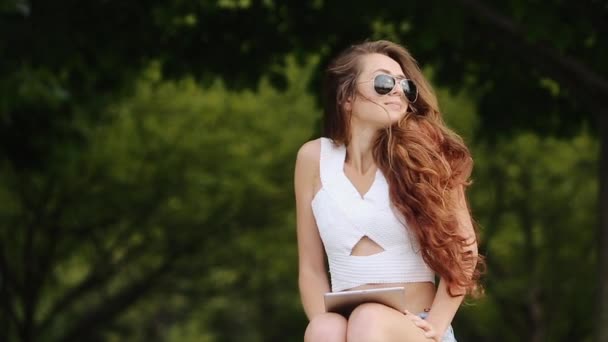 Menina bonito em camisa branca e Jean Shorts em sentado no banco com seu dispositivo e árvores verdes no fundo — Vídeo de Stock