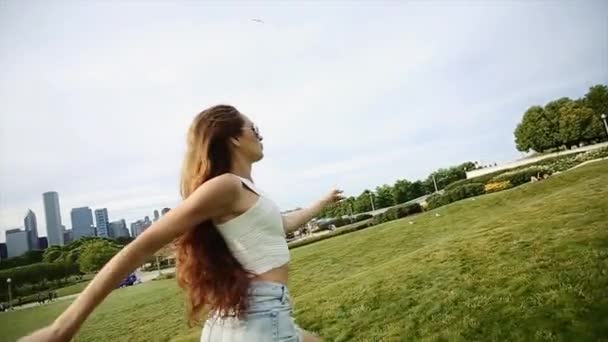 Uma menina com longos cabelos castanhos vestindo uma camisa branca, calções Jean, sapatos brancos e óculos de sol pretos está correndo em um parque . — Vídeo de Stock