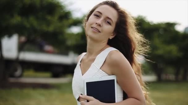 Une fille attrayante avec un sourire radieux joue avec ses cheveux comme elle se déplace autour tenant une tablette sur une journée ensoleillée lumineuse . — Video