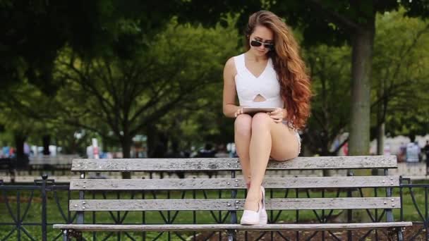 Linda chica de camisa blanca y pantalones cortos de Jean en sentarse en el banco con su dispositivo y la gente y los árboles verdes en el fondo — Vídeos de Stock