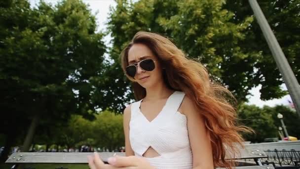 Belle fille en chemise blanche et lunettes de soleil noires est assis sur le banc et parler au téléphone avec des arbres verts en arrière-plan au ralenti — Video