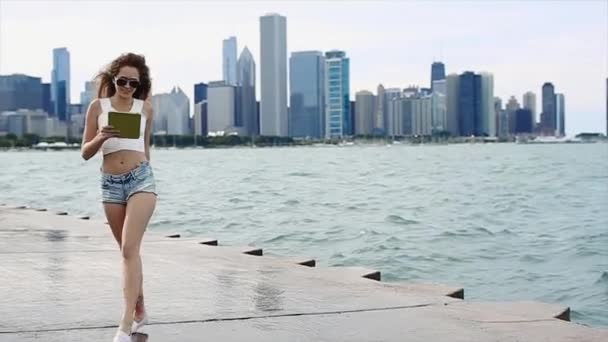 Schönes Mädchen mit langen, lockigen Haaren in weißem Top und Jeanshose geht in Zeitlupe mit Gerät in den Händen am See entlang — Stockvideo