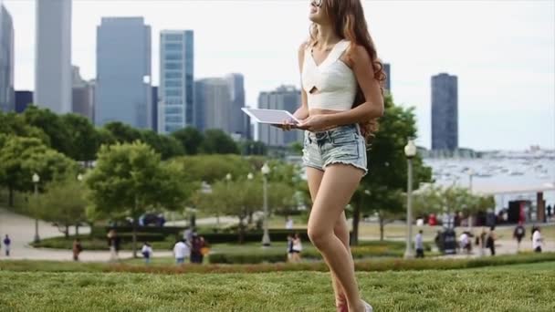 Pretty Caucasian Girl en camisa blanca y pantalones cortos Jean está jugando con el dispositivo con edificios altos, lago y árboles en el fondo en cámara lenta — Vídeo de stock