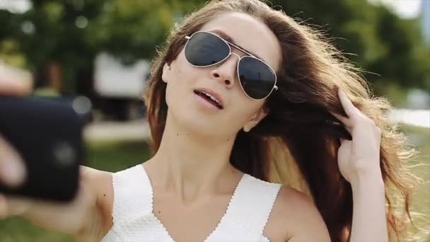 Menina bonita em camisa branca e óculos de sol pretos tomando um selfies com lago e edifícios altos no fundo — Vídeo de Stock