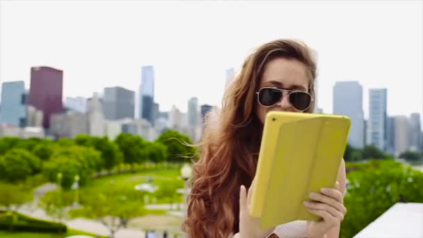 Jolie caucasienne en chemise blanche et lunettes de soleil noires aux cheveux longs parle sur son appareil avec de grands bâtiments en arrière-plan au ralenti — Video